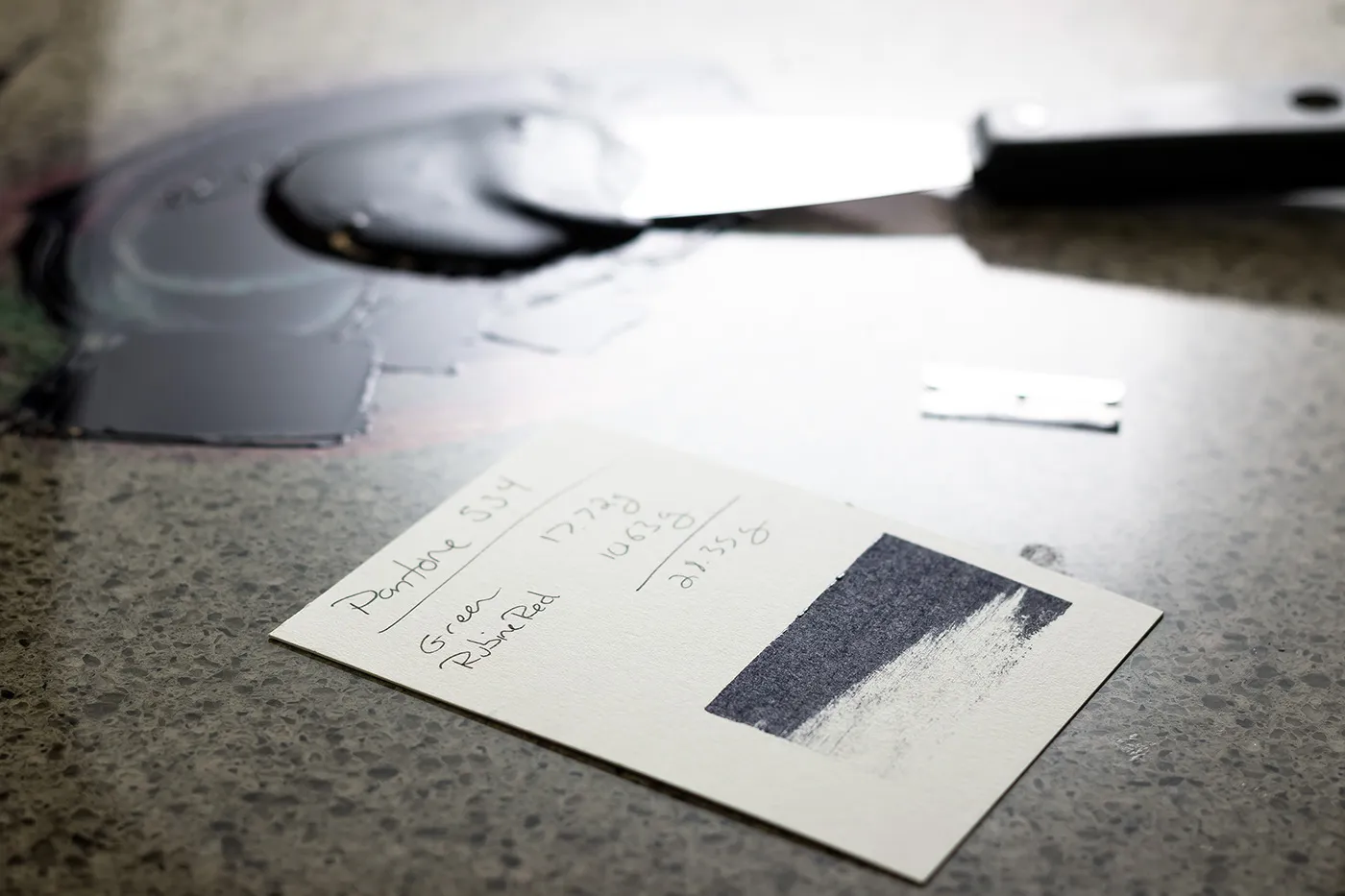 Handwritten Pantone formula card with a hand-mixed ink drawdown swatch beside the ink palette and knife in the studio